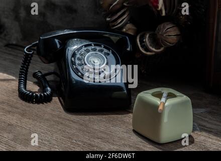 A litted cigarette on greyish green ashtray which is placed next to old-fashion telephone. The heart-shape wreath as background. Selective focus. Stock Photo