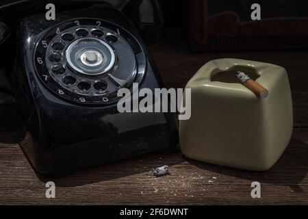 A litted cigarette on greyish green ashtray which is placed next to old-fashion telephone. Selective focus. Stock Photo