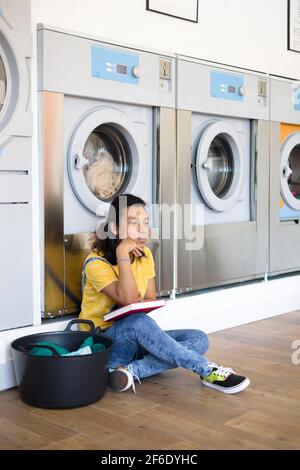 Young latin american woman bored while waiting for clothes washing at the self-service laundry. She has a book in her hands. Stock Photo