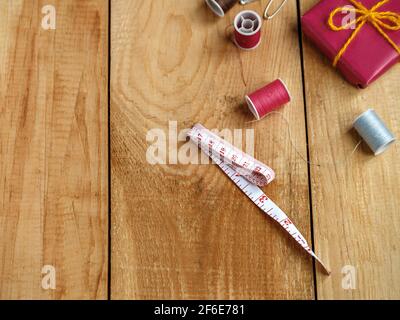 Tailor's measuring tape, thread coils and red gift box on brown wooden boards. Stock Photo