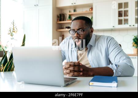 Shocked amazed African American man with glasses, freelancer, ceo or stock agent sits at his desk, looking at laptop in surprise, reading unexpected news or got big account profit Stock Photo