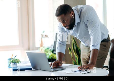 Busy influential serious African American male entrepreneur or top manager, in formal wear, stands near a work desk, uses a laptop, looks for information, responds to email, browsing internet Stock Photo