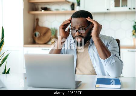 Worried African American man with glasses, sits at his desk, looking at laptop in confusion, holding his head with his hands, reading bad news, received an unexpected e-mail message, failed the deal Stock Photo