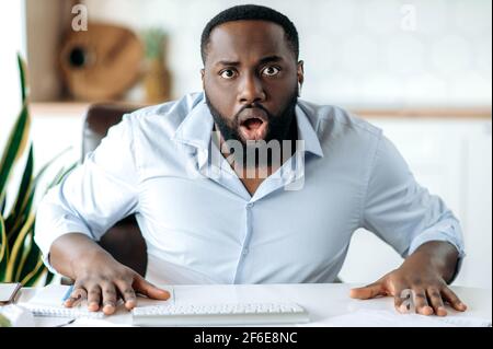 A surprised shocked African American bearded guy wearing a formal shirt, received unexpected news, amazed looking at the camera while sitting at the workplace Stock Photo
