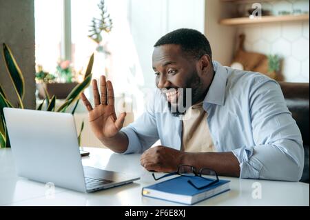 Virtual online meeting. Joyful African American young stylish man, sits at work desk, uses laptop to communicate by video call with colleagues or business partners, greeting with hand gesture, smiling Stock Photo