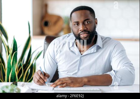 Handsome friendly African American young man, businessman, expert, assistant or broker, wearing formal blue shirt, sitting at work desk, taking notes, looking at camera, smiling Stock Photo