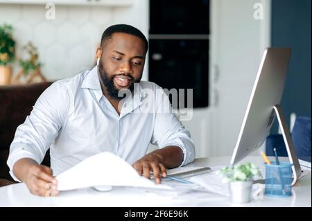 Busy successful African American man, entrepreneur or lawyer, in formal wear, sits at work desk, uses computer for work, studies financial documents, analysis profit and strategy of company Stock Photo