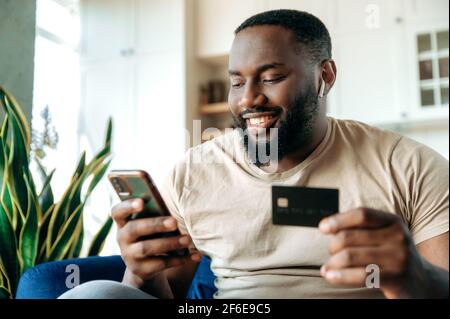 A satisfied smiling african american guy, in casual clothes, sits on the couch at home, holds a phone and a credit card in hands, pays for online purchases, enters a card number, online shopping Stock Photo