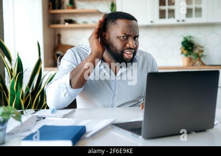 Afro american confused male freelancer or manager holding hand near ear, showing gesture of listening, while sitting at work desk and laptop, is listening news Stock Photo