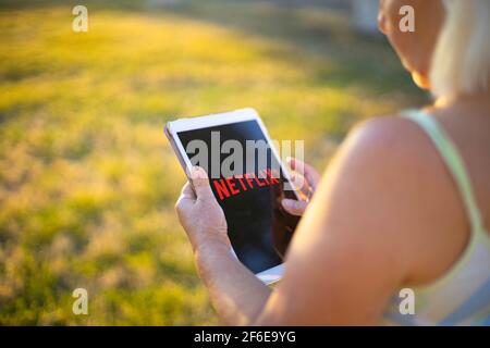 Podolsk, Ukraine - March 29, 2021: Woman watches Netflix sitting and holding tablet on green grass at the park Stock Photo