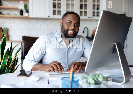A portrait of an attractive good looking african american successful guy, real estate agent, lawyer or ceo, manager, wearing formal shirt, sits at workplace at home, looks at camera, friendly smile Stock Photo