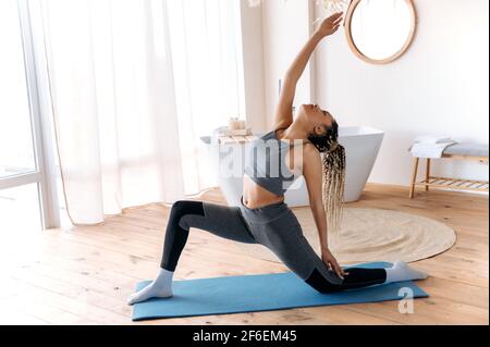 Sporty beautiful African American young woman with good figure, in fitness wear and with dreadlocks, leads a healthy lifestyle, takes care health, does fitness and stretching at home on the rug Stock Photo