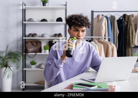 Young african american showroom owner holding thermo cup and using laptop near notebooks and smartphone Stock Photo