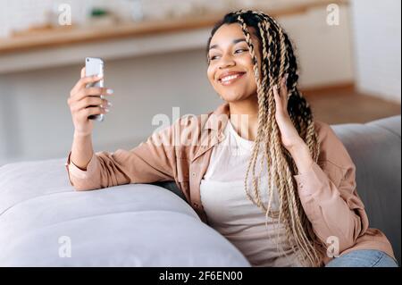 Cute happy african american stylish girl with dreadlocks at home, communicates on video conference using smartphone, does selfie, gestures with hand, smiling happily Stock Photo