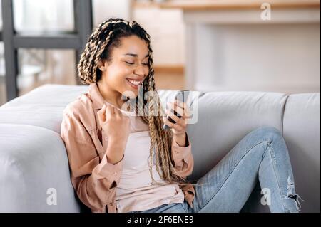 Joyful attractive african american girl with dreadlocks, sitting on the couch, using smartphone, rejoices success or win, get a discount for shopping, gestures with her hand, smiles Stock Photo