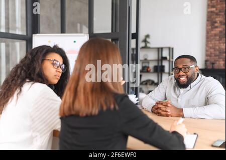 The working process at contemporary coworking. Diverse team of young ambitious people brainstorming together, an African-American male in eyeglasses listening carefully his female colleague Stock Photo