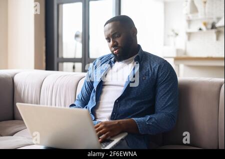 African-American guy in casual wear using the laptop, spends leisure time online in network sitting on the comfortable couch, black man working remotely Stock Photo