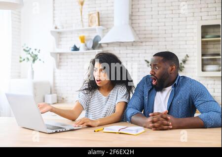 Shocked multiracial couple staring on the laptop screen, saw unexpected bad news or received huge utility bills by email. Biracial woman and African man looks at computer with mouths open surprised Stock Photo