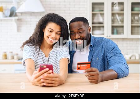 Multiracial couple sits in the kitchen at home and making online order of food, using smartphone app for purchasing online, enjoys shopping together Stock Photo