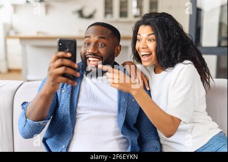 No way. Surprised multiracial couple looks at the smartphone screen with astonishment, African guy holds phone, charming biracial girlfriend points finger at screen, watching funny movies and laughing Stock Photo