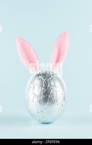 Easter egg wrapped in a silver foil with pink bunny ears against pastel blue background. Easter minimal concept. Creative Happy Easter or spring layou Stock Photo