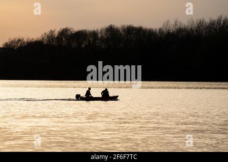 people in speed boat silhouette at sunset on lake with golden light reflecting on river water sailing quickly to rescue swimmers. Reservoir calm water Stock Photo