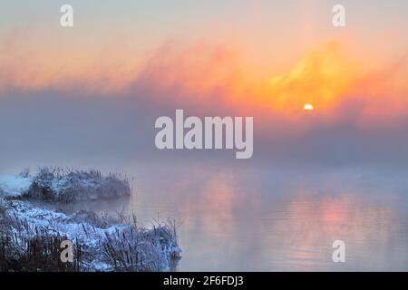 Sunrise time over the winter river. An orange fog spreads over the water and obscures the horizon. Reeds stand in the snow near the water. Stock Photo