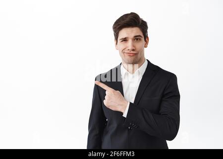 Displeased boss, businessman in black suit frowning and shaking head upset with bad result, pointing left with skeptical face, standing disappointed Stock Photo