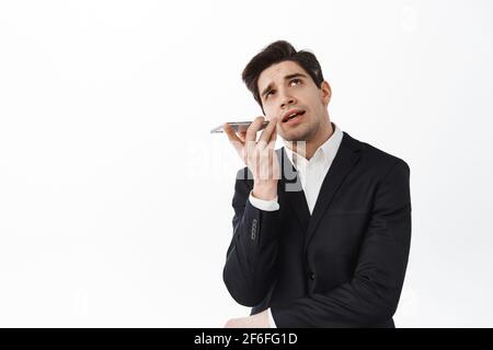 Businessman talking on speakerphone, look up thoughtful and record voice on smartphone, using translator app on cellphone, standing over white Stock Photo