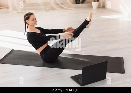 Woman trainer practicing yoga performs the Naukasana exercise, boat pose, showing the correct execution to the students using a laptop. Concept on the Stock Photo