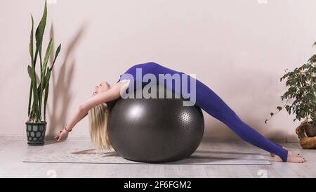 Attractive woman in blue sportswear practices yoga, performing bridge pose, bandhasana on fitball in the room near the wall. Stock Photo