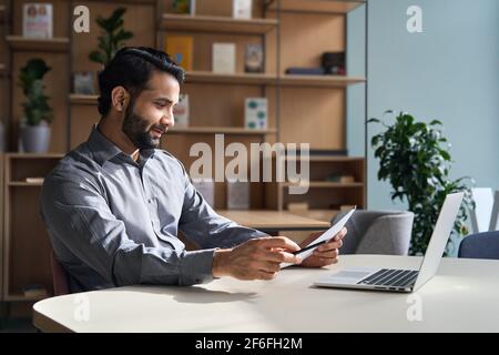 Smiling indian business man working on laptop checking document at home office. Stock Photo
