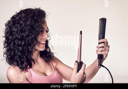 Young smiling curly-haired woman in profile holds a hair curler and a hair straightener. care and beauty concept Stock Photo