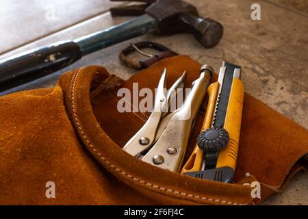 Home improvement tools in a camel colored brown suede tool belt. A multitool, a hammer, and a precision box-cutter knife with yellow edges. Soft-focus. Stock Photo