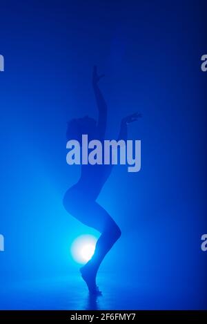Ballerina in black bodysuit dancing with magic blue light and smoke. Silhouette of young attractive dancer in ballet shoes pointe performing in dark. Stock Photo