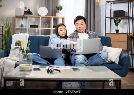 Group of two young people using personal laptops while doing homework. Asian students sitting on couch together and studying. Concept of technology and education. Stock Photo