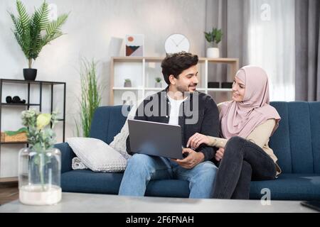 Relaxed Arabian Muslim couple in casual wear, using a laptop pc together for watching online content or shopping, sitting on a sofa in the living room at home. Stock Photo
