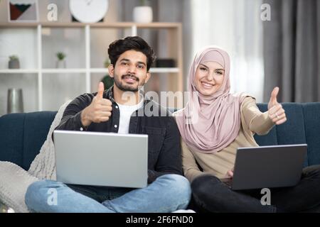 Young multiracial Muslim couple relaxing on sofa at home using laptops, smiling at camera and showing thumbs up. Stock Photo