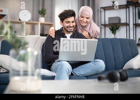 Young handsome bearded arabian man sitting on the sofa and watching movie or news on laptop at home, while his pretty smiling wife in hijab, standing behind him and looking at the screen. Stock Photo