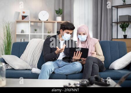 Coronavirus concept, stay at home. Young arabic couple in medical masks, sitting together at home on sofa and using tablet pc to read news or social networks, discussing them. Stock Photo