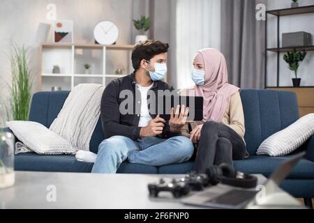 Couple in masks sitting on sofa at home. Young Arabian Muslim couple, spending time during desease and isolation at home, using digital tablet for watching movie or byuing food online Stock Photo