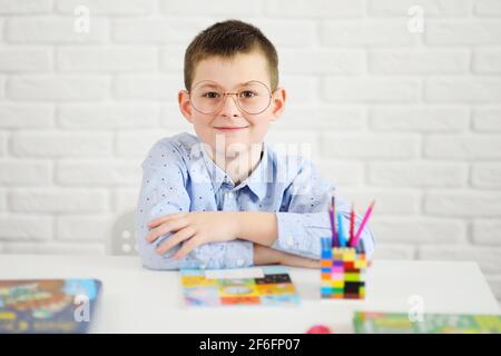 a schoolboy with glasses sitting at a white table smiles against the background of notebooks and books. Stock Photo
