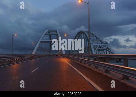 Cars go under the arches of the Crimean bridge in the late evening. On the right, a railway bridge is being built. Dull Stock Photo