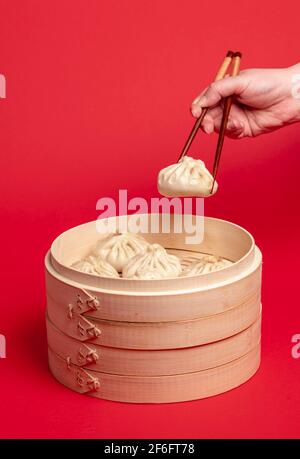Woman taking a dumpling from the bamboo steamer with the chopsticks. Freshly steamed baozi dumplings in a wooden steamer isolated on a red colored bac Stock Photo