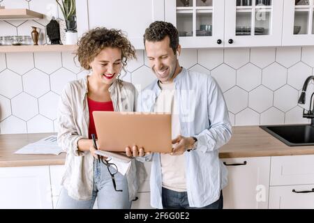 Happy family couple are standing at the kitchen at home, doing online shopping or chatting with friends. Joyful stylish husband and wife using laptop Stock Photo