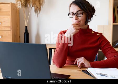 Woman wearing red turtleneck and eyeglasses works with laptop near the window. Remote working concept. Stock Photo