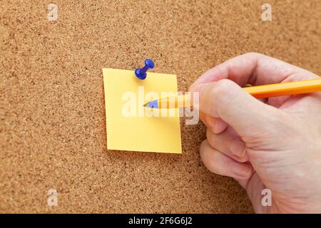 A man writing on a paper note with a ballpoint pen that is pinned on a cork board. Close up. Stock Photo
