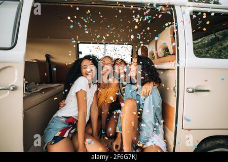 Group of happy women sitting in a camper van door under confetti. Friends having fun Stock Photo