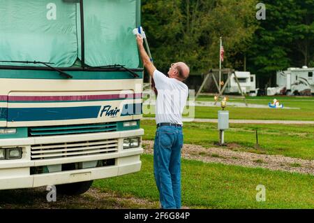 An elderly man cleans the windshield of his older class A Flair RV camper van motor home in a campground. USA. Stock Photo