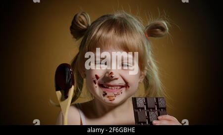 Joyful smiling child kid girl with dirty face from melted chocolate on dark background in studio Stock Photo